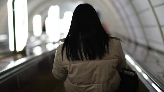 Portrait of woman with dark hair and bob haircut in warm jacket descending escalator in subway back view

