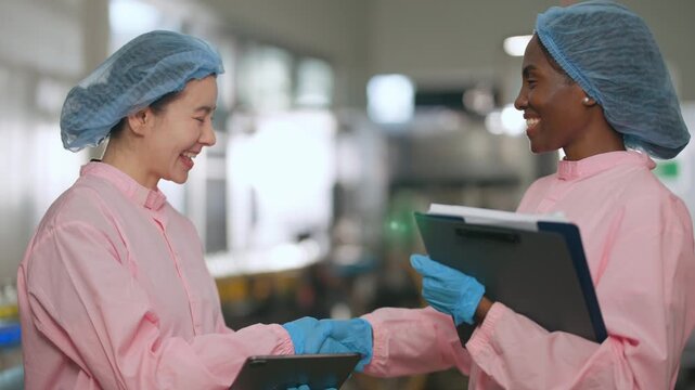 Two Women in Lab Coats Collaborating: Two female scientists, one Asian and one Black, wearing pink lab coats and hairnets, smile and collaborate in a laboratory setting.