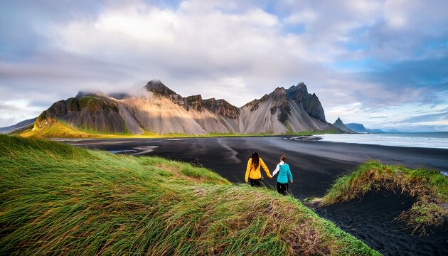 two adventurers walk hand in hand on the striking black sand beach of vestrahorn surrounded by dramatic mountain peaks and vibrant green grass stokksnes beach vestrahorn mountain iceland