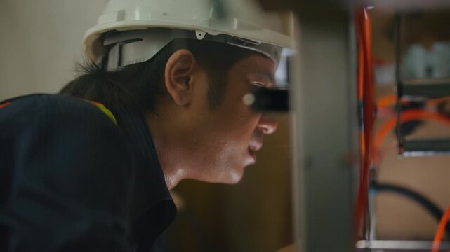 Focused Electrician at Work: A diligent electrician wearing a hard hat and work uniform intently examines electrical wiring and equipment.