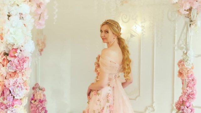 fantasy happy girl princess in white room blooming spring scenery, flower decor arches rose. Woman queen blonde hair, vintage pink evening dress back view. Cheerful lady looking back smiling face. 4k