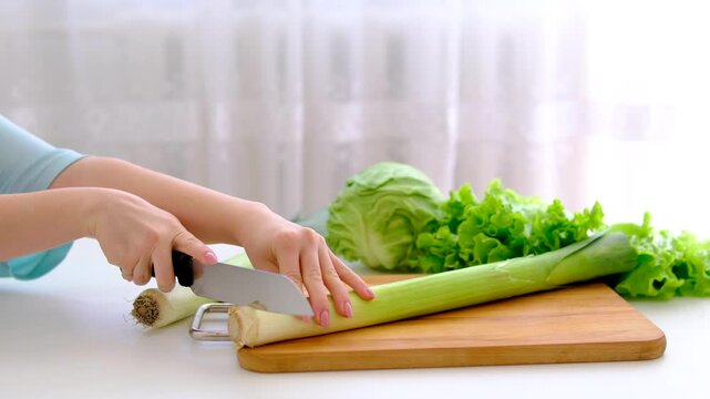Slicing fresh leek on wooden board in bright white kitchen with cabbage and lettuce