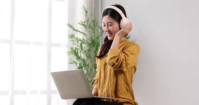 Young asian woman smiling and listening to music through headphones while using a laptop for studying or remote work, creating a focused and joyful atmosphere in her home office setup
