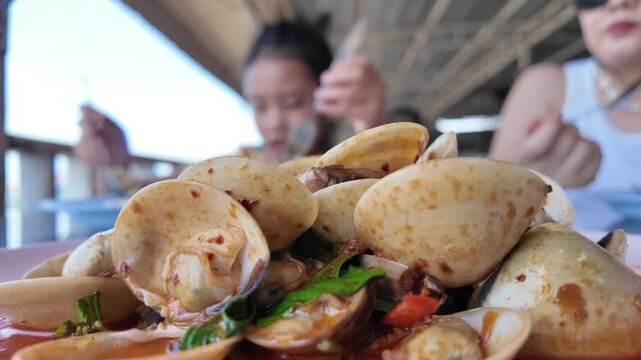 A Delicious Meal: A close-up shot of a plate filled with delectable shellfish, glistening in a savory sauce, with people enjoying a meal in the blurred background.