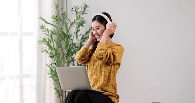 Young asian woman smiling and listening to music through headphones while using a laptop for studying or remote work, creating a focused and joyful atmosphere in her home office setup