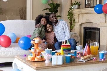 Fototapeta premium African American family with child smiling, hugging on couch at home with rainbow cake, balloons