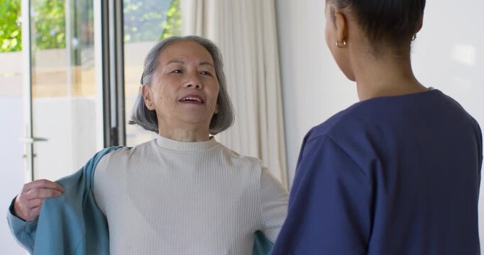 Caregiver helping senior Asian woman lifting blue jacket, adjusting collar at sliding glass door