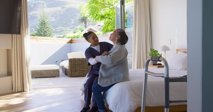 African American female carer steadying Senior Asian woman leaning, helping settle on bed by walker