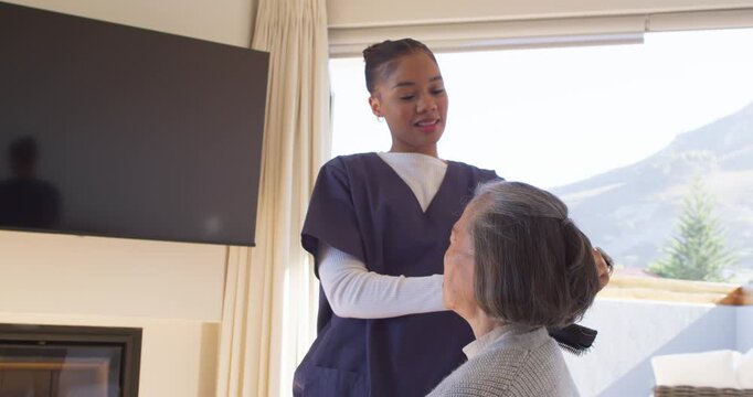 African American aide scrub-clad greeting styling Asian senior into bun for care with comb at home
