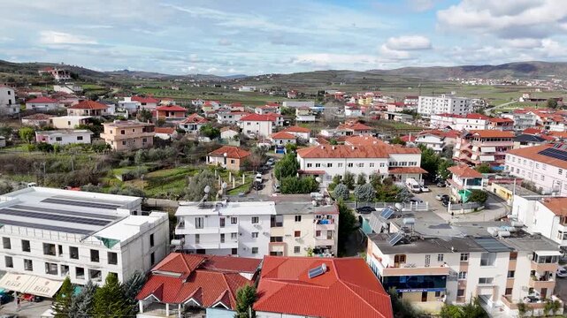 Wide aerial tracking shot pulling back over the calm dark surface of Lake Belsh, revealing dense red-roofed town apartments, lakeside promenades, and terraced agricultural hills in Albania