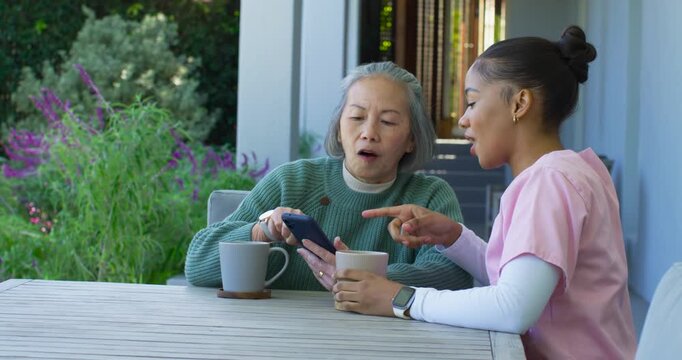 Senior Asian woman holding phone and female caregiver in pink scrubs pointing, guiding her on porch