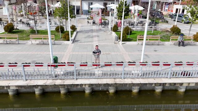 Aerial tracking shot pulling back and rising from host Charles Huang of DIY Destinations to reveal a lakeside pedestrian plaza, the calm waters of Lake Belsh, and distant rolling hills