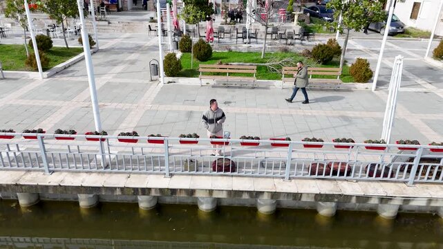 Aerial tracking shot pulling back and rising from host Charles Huang of DIY Destinations to reveal a lakeside pedestrian plaza, the calm waters of Lake Belsh, and distant rolling hills
