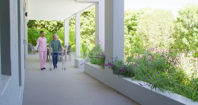 African American carer in scrubs pointing Asian senior with walker past plants on porch, copy space