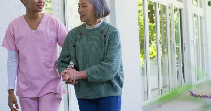 African American nurse pink scrubs meeting Asian senior at glass doors holding hand walking for aid