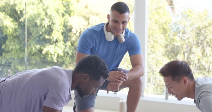 Male trainer checking watch, starting two trainees performing synced pushups on blue mats in gym