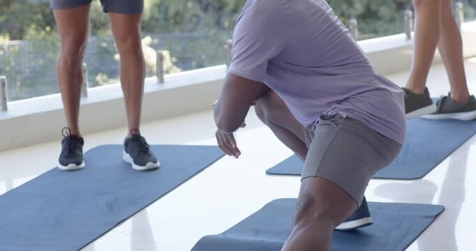 Diverse male group lunging and squatting on blue mats in bright studio, leader checking smartwatch