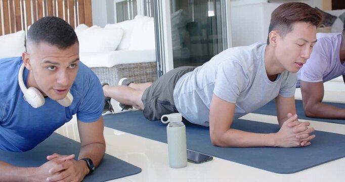 Diverse male friends after warmup, holding forearm planks at home with water bottle, phone for core