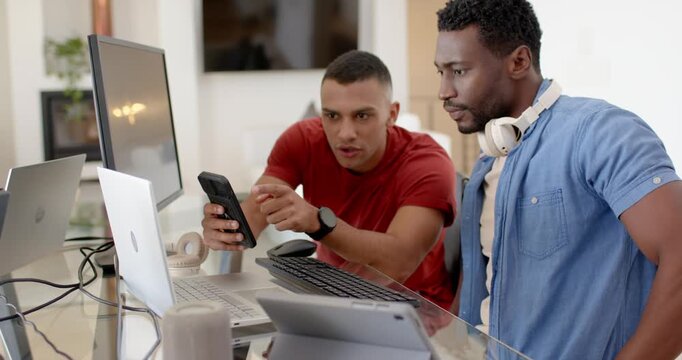 Two male coworkers at desk, red tee, blue shirt headset showing phone to monitor, typing, checking