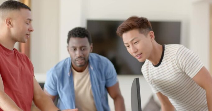 Diverse male friends crowding at table, red-shirt gesturing toward tablet, celebrating win