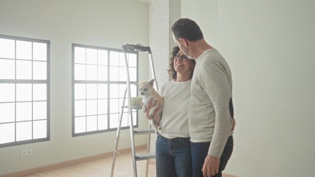 Man and woman embrace while holding a small dog beside a step ladder in building; new home contentment.
