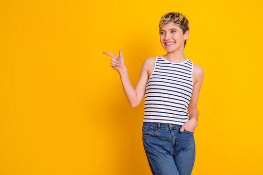 Young smiling woman pointing to the side on a bright yellow background wearing a striped top and jeans in a casual confident pose