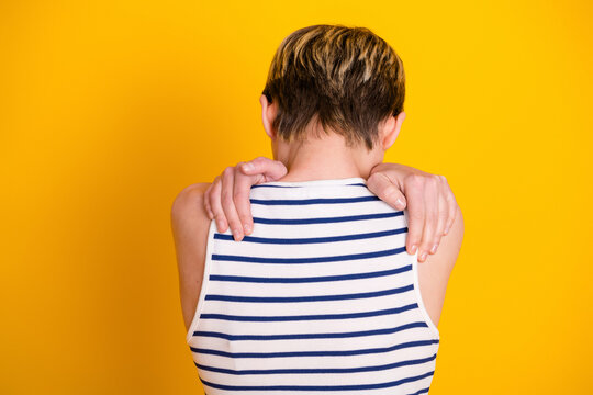 Young person with short hair wearing a striped tank top holding their shoulders in a self-hug gesture against a bright yellow background