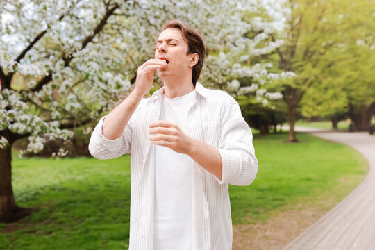 A man suffering from seasonal allergy and hay fever sneezes in a nature park surrounded by blooming spring trees with pollen.