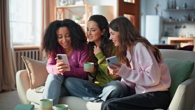 Three besties browsing phones sitting together in living room. Diverse friends