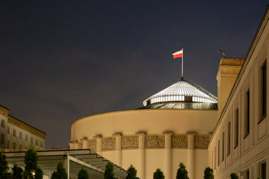 Night view of the illuminated dome of the Polish Parliament Sejm building in Warsaw featuring bas-reliefs and the national flag of Poland.