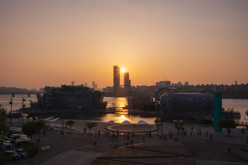 Naklejka premium Golden sunset over the Banpo Hangang Park paints the Hangang skyline with warm light, while the nearby Moonlight Rainbow Fountain adds vibrant energy to the evening cityscape.