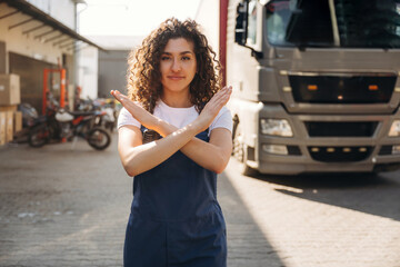 Naklejka premium Young woman with curly hair in blue overalls stands with arms crossed in front of a large truck in a loading area, showcasing a confident stance