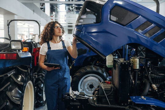 Female mechanic in blue overalls inspects tractor engine with tablet in hand, surrounded by agricultural machinery in a well-lit workshop setting