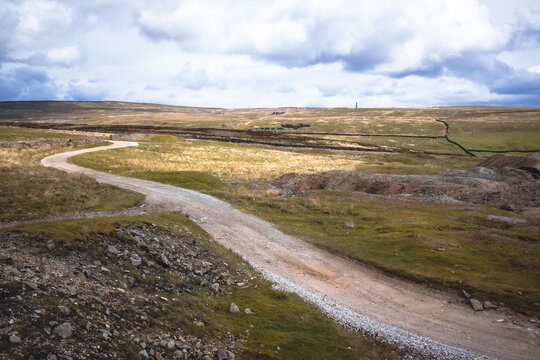 Winding gravel track passing historical lead-mining ruins on Grassington Moor in the Yorkshire Dales, featuring the iconic Cupola Smelt Mill Chimney under a dramatic cloudy sky.