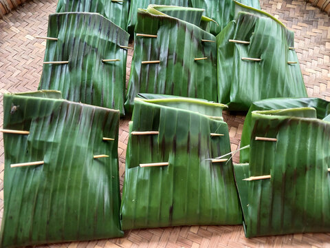 Traditional Indonesian food wrapped in fresh green banana leaves secured with toothpicks. Authentic organic packaging on woven bamboo background close-up view.