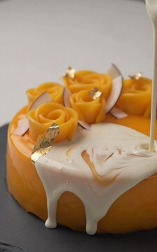 A close-up view of a round orange cake with white icing and mango roses on top.