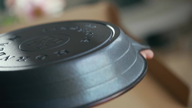 Hand lifts heavy black cast iron skillet. Smooth, rounded edges and embossed logo visible. Skillet rests on cardboard box. Action shows careful handling of cooking tool. Ideal for food, cooking
