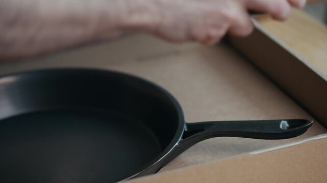 Hand carefully removes a black cast iron skillet from a cardboard box. The skillet is smooth and ready for use. Close-up shot shows the object and hand in detail. Ideal for cooking, kitchen
