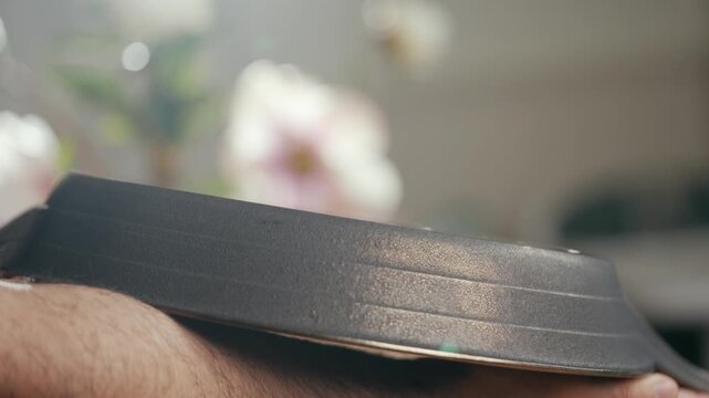 Hand holds black frying pan close-up. Smooth metal surface reflects soft light. Background blurred with floral decor. Focus on cooking tool detail and texture. Ideal for culinary, kitchen