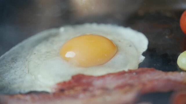 Egg drops onto hot skillet, sizzles, and cooks beside crispy bacon. Smoke rises as yolk settles perfectly. Close-up shot captures cooking texture and warmth. Ideal for breakfast, cooking tutorials