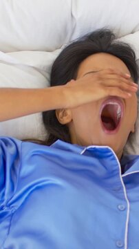 Vertical video: Yawning woman in blue pajamas covering mouth then rubbing eyes while waking in bed
