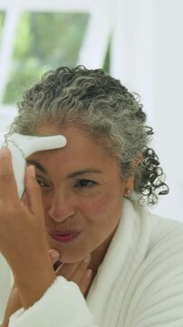 Vertical video: Pressing device woman in robe at vanity moving from cheek to brow forehead for skin