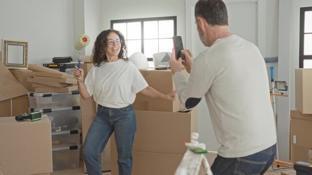 Woman holding a paint roller and smartphone, smiling and dancing amid moving boxes in a building during home renovation; joy teamwork.