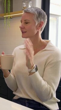 Vertical video: Holding mug, woman in cream sweater offering mug in kitchen as friend entering left