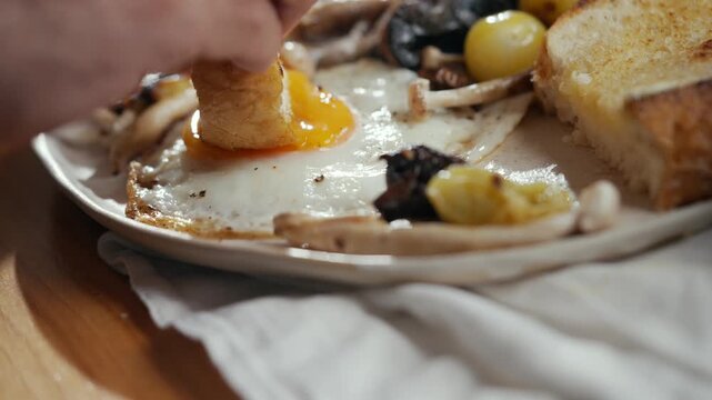 Hand dips bread into sunny-side-up egg. Golden yolk glistens as bread absorbs flavor. Toast and olives surround plate. Warm, cozy breakfast moment. Perfect for food blogs, recipes, or morning scenes