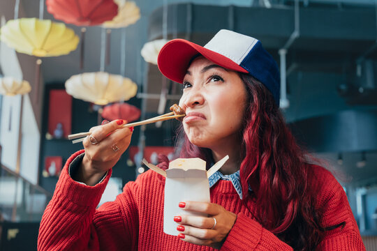 Young woman with red hair wearing a cap, expressing disgust while trying a piece of Asian food from a takeout box