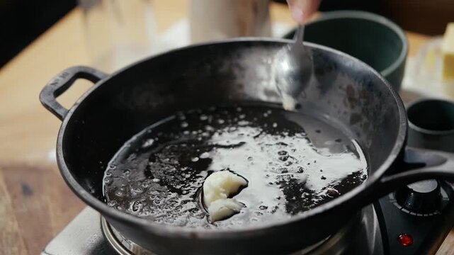 Hand drops garlic into hot oil in black pan. Oil bubbles around garlic as it fries. Close-up on cooking process in rustic kitchen. Scene suggests home cooking or meal prep. Ideal for culinary, food