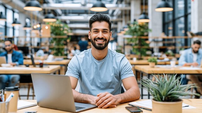 Smiling bearded middle eastern man sitting at wooden desk with laptop in modern open coworking office space and looking directly at camera