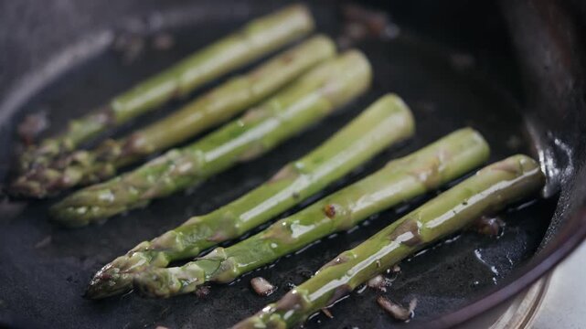 Fresh green asparagus spears sizzle in a dark frying pan. Oil glistens around the spears as they cook. Tiny bits of garlic or onion add flavor. Simple, rustic cooking scene with no people shown