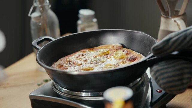 Close-up of hands lifting a cast-iron skillet with freshly baked pizza. Skillet sits atop a portable stove on wooden countertop. Background contains kitchen items like bottles and jars
